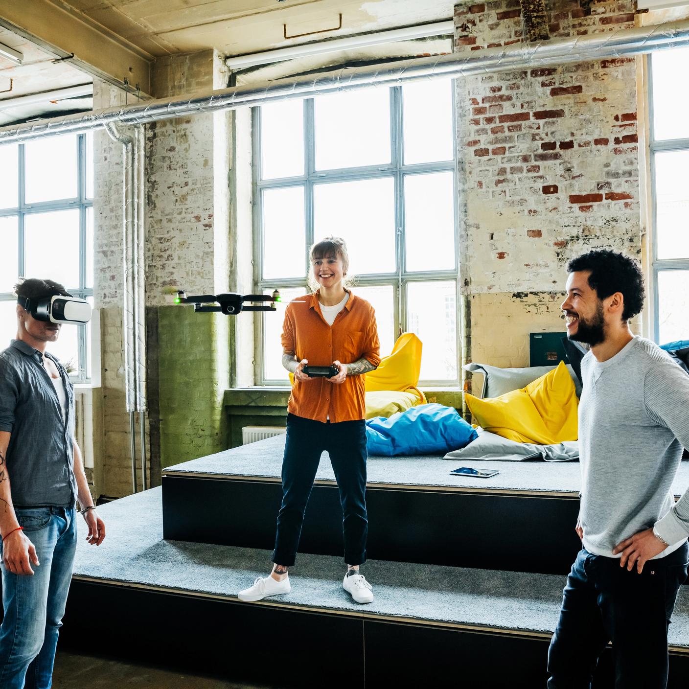 Young start up business people flying a drone inside a big office. One man wearing VR googles and testing new augmented reality software with the drone camera