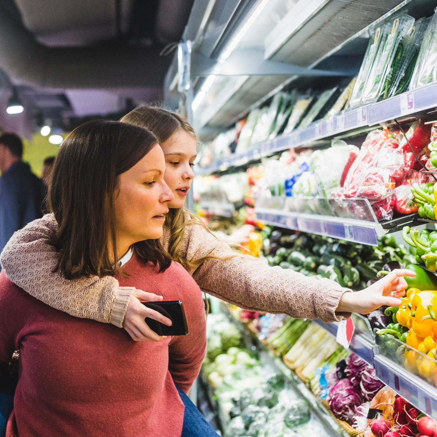 Daughter choosing bell peppers while being piggybacked by mother in store