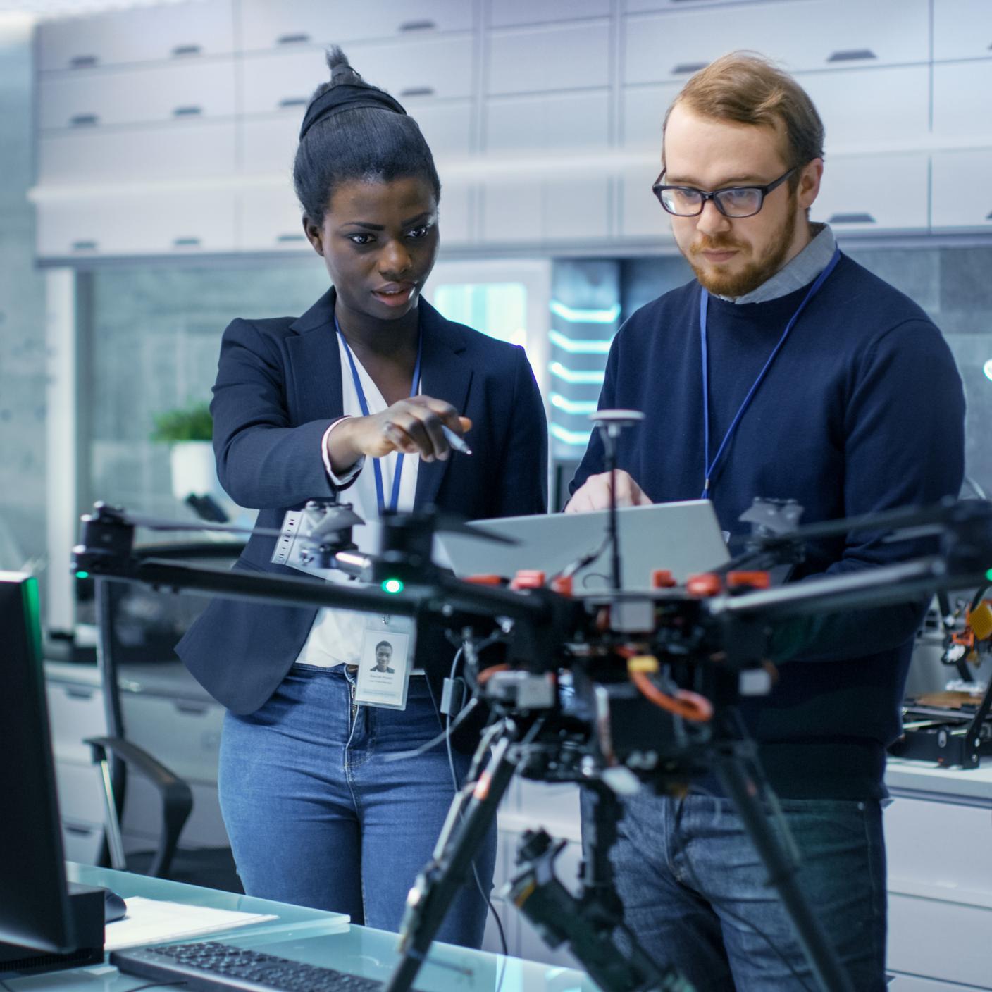 Caucasian Male and Black Female Engineers Working on a Drone Project with Help of Laptop and Taking Notes.