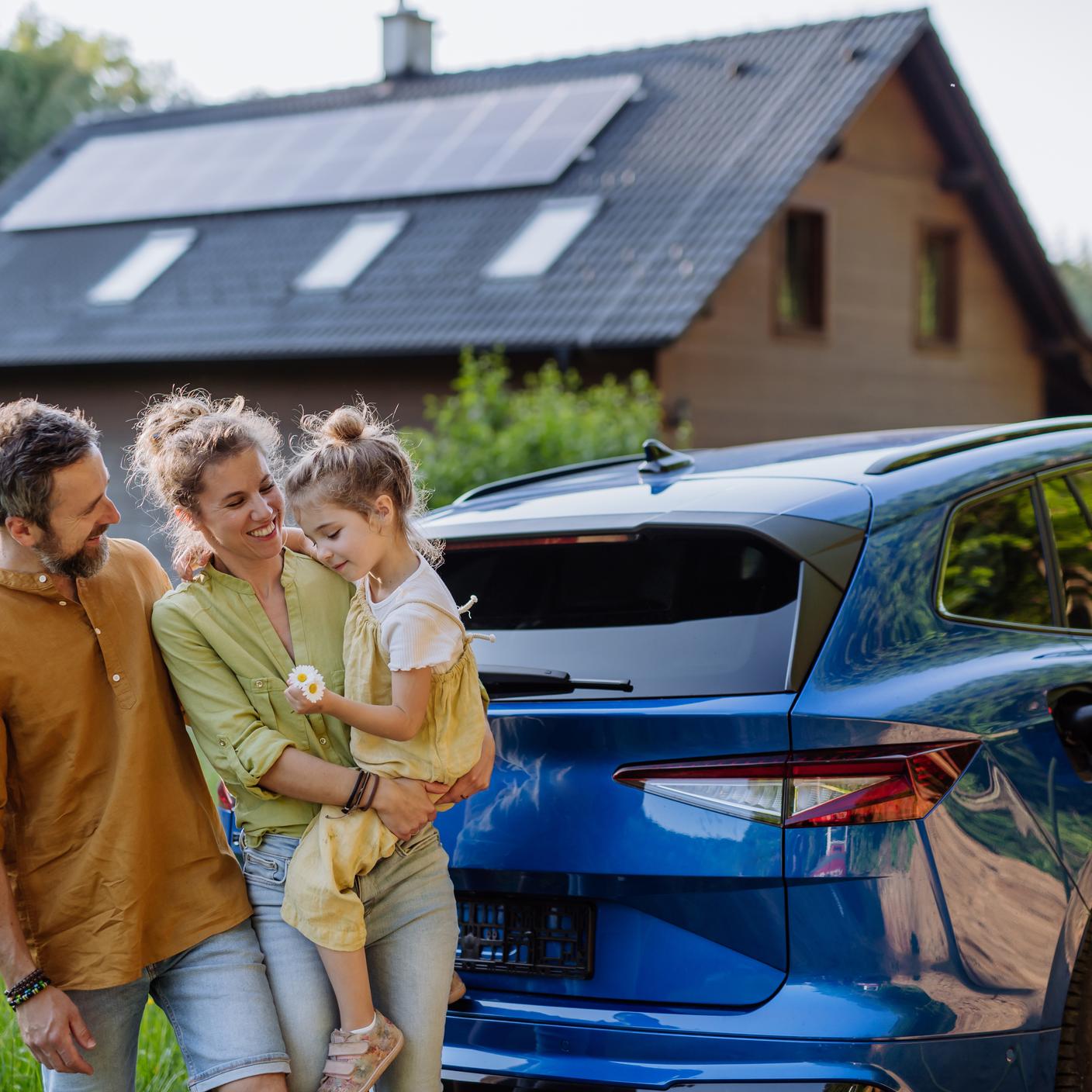 Family with little daughter standing in front of their house and electric car