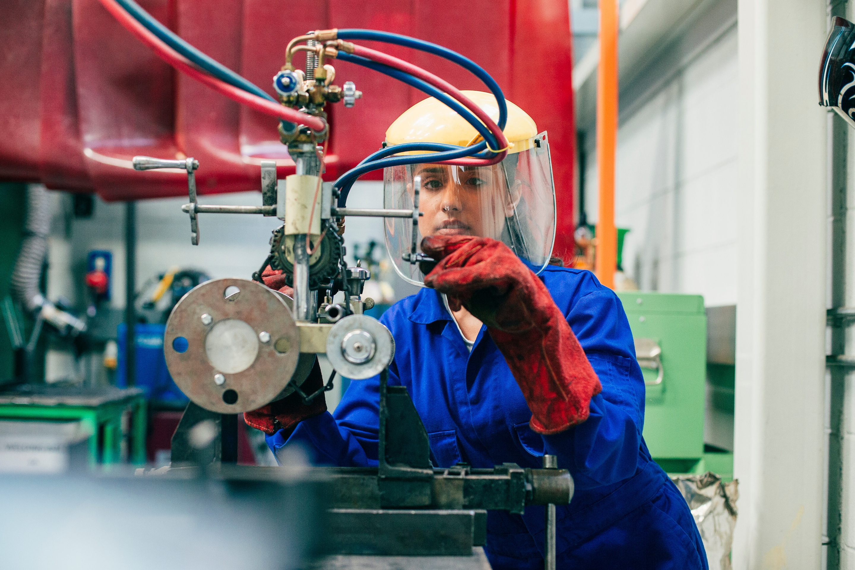 A young woman is using engineering equipment in a workshop. She is wearing a protective mask and gloves as well as blue coveralls.