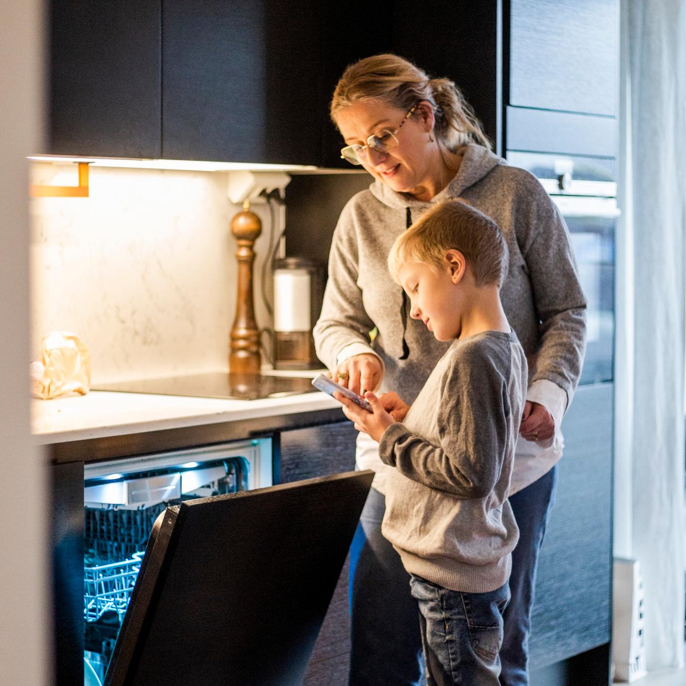 Une mère apprend à son fils à utiliser une application mobile tout en faisant fonctionner le lave-vaisselle dans la cuisine à la maison