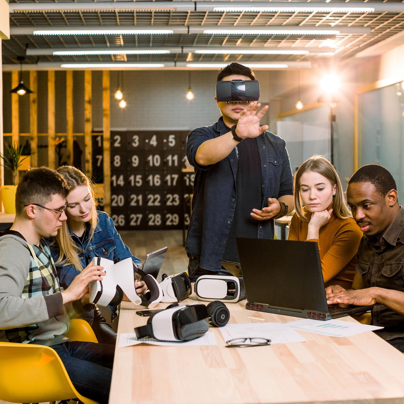 Teamwork trying virtual reality glasses for work in creative office. Young Asian man using new technology vr goggles while his coworkers talking sitting at the table