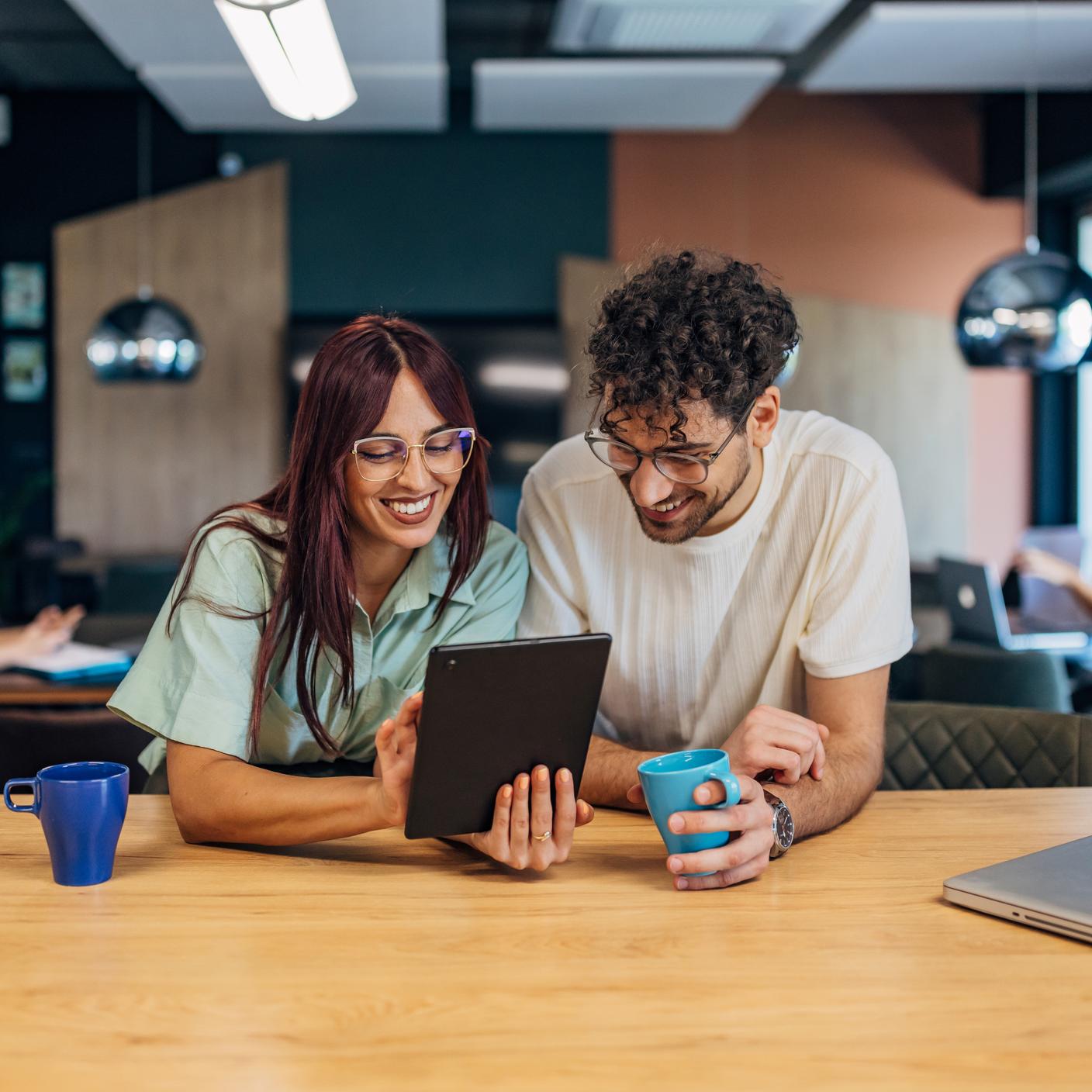 Two cheerful business colleagues drink coffee and use a digital tablet while designing a business project they are working on as a team.