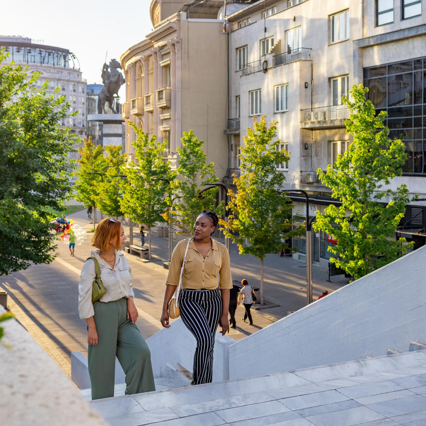 Two female friends having a walk in an urban city walking street, surrounded by people, modern buildings and greenery.