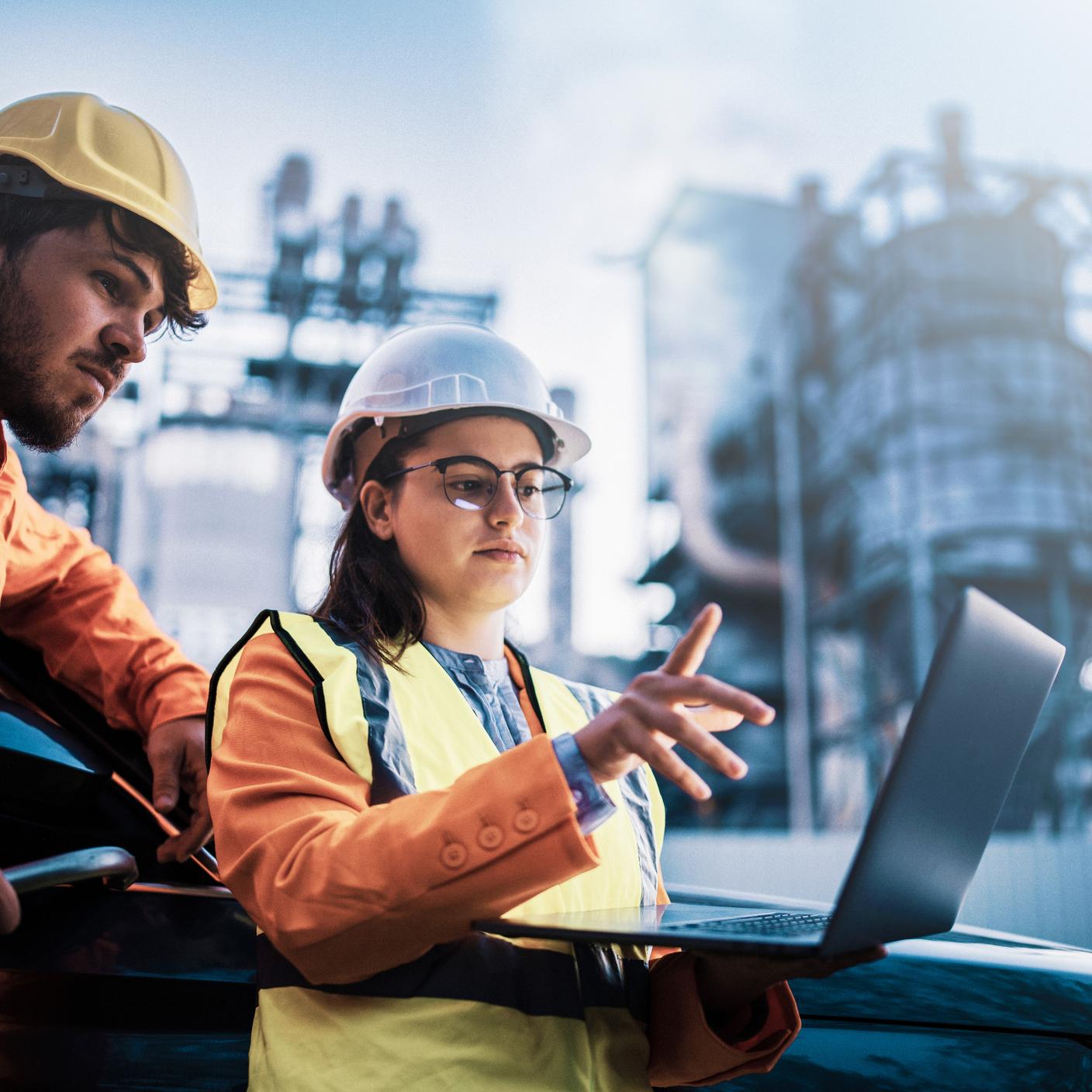 Two associates in safety attire reviewing data on a laptop in front of a power plant.