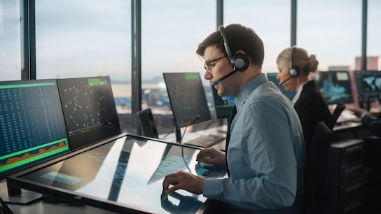 Male Air Traffic Controller with Headset Talk on a Call in Airport Tower. Office Room is Full of Desktop Computer.