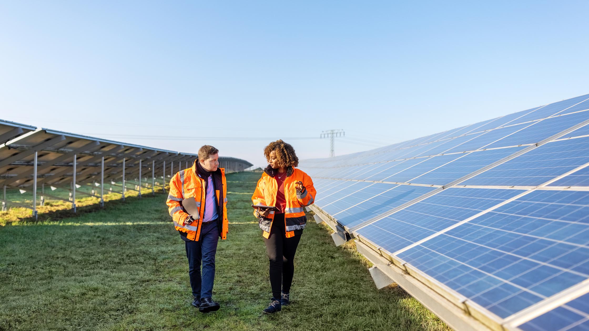 Two technicians in reflective clothing walking between rows of photovoltaic panels at solar farm.
