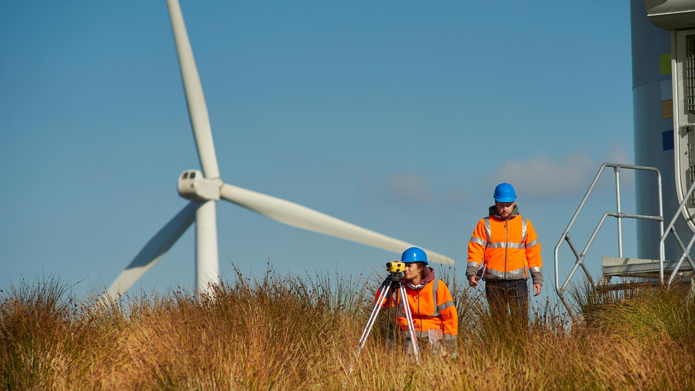 two wind farm engineers leaving the control panel of a wind turbine. They are assessing their readings on a digital tablet as they walk away. In the foreground a theodolite can be seen .