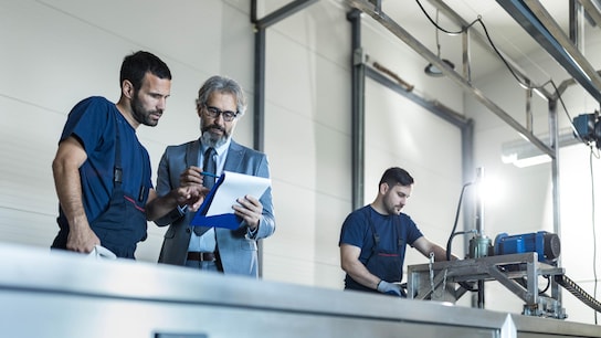 Mature inspector and manual worker going through paperwork in a factory. Other worker is in the background.