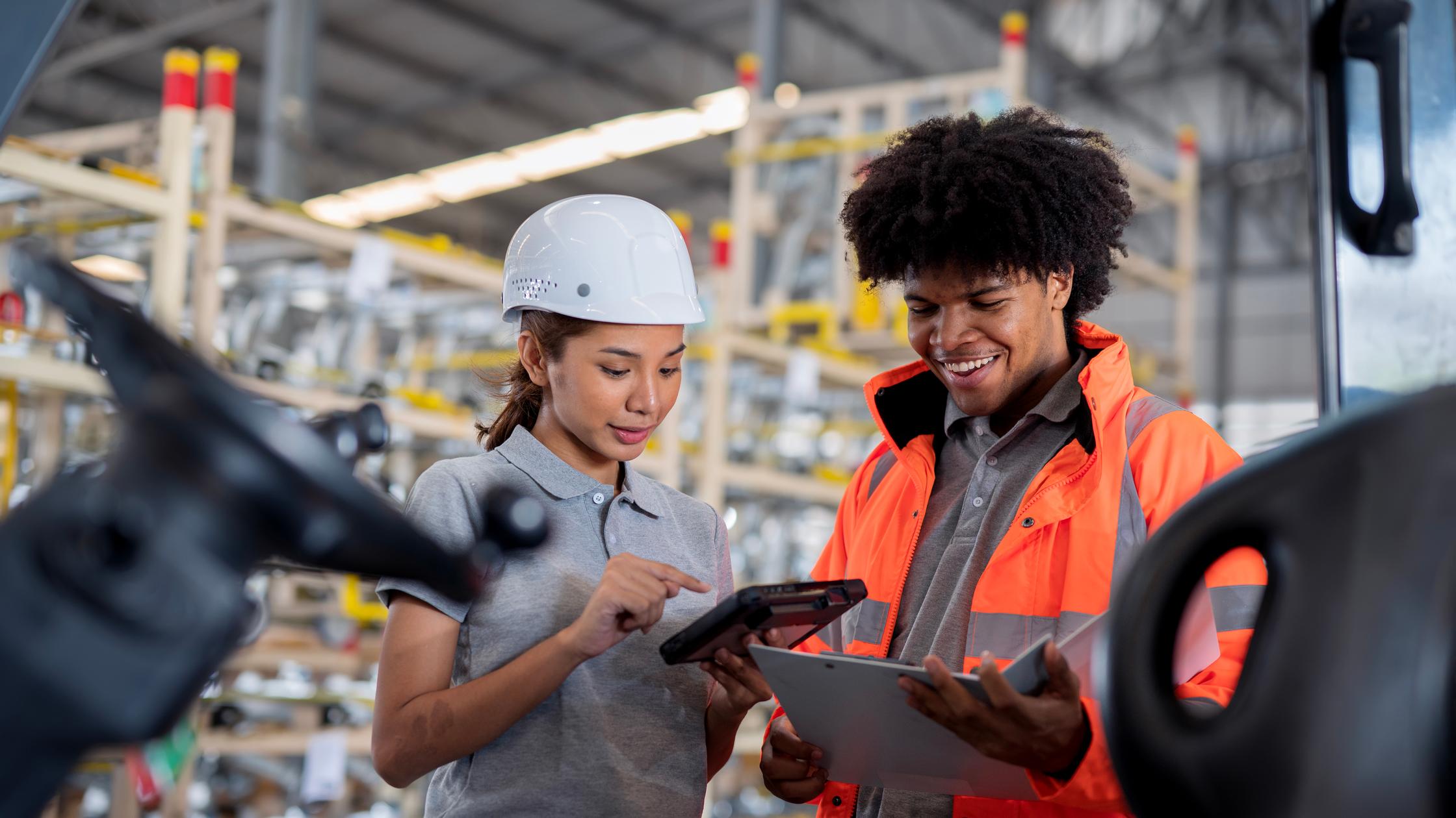 A Warehouse Supervisor discussing a tablet computer with warehouse workers behind a forklift about delivery schedule in a factory warehouse.