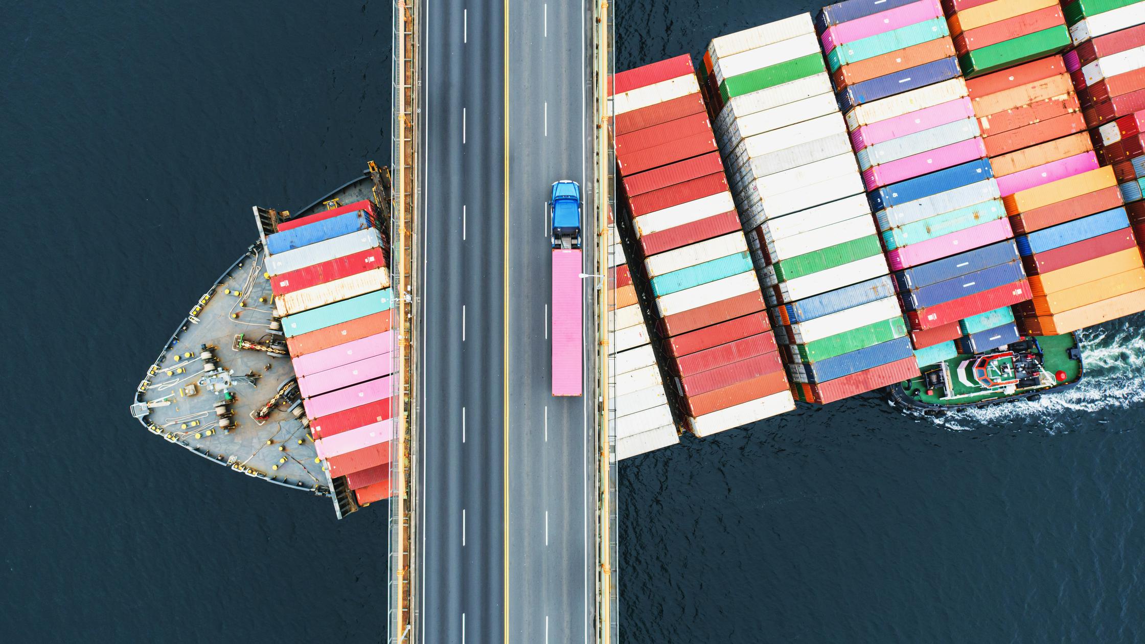 Aerial view of a container ship passing beneath a suspension bridge. Semi truck with pink cargo container crosses above.