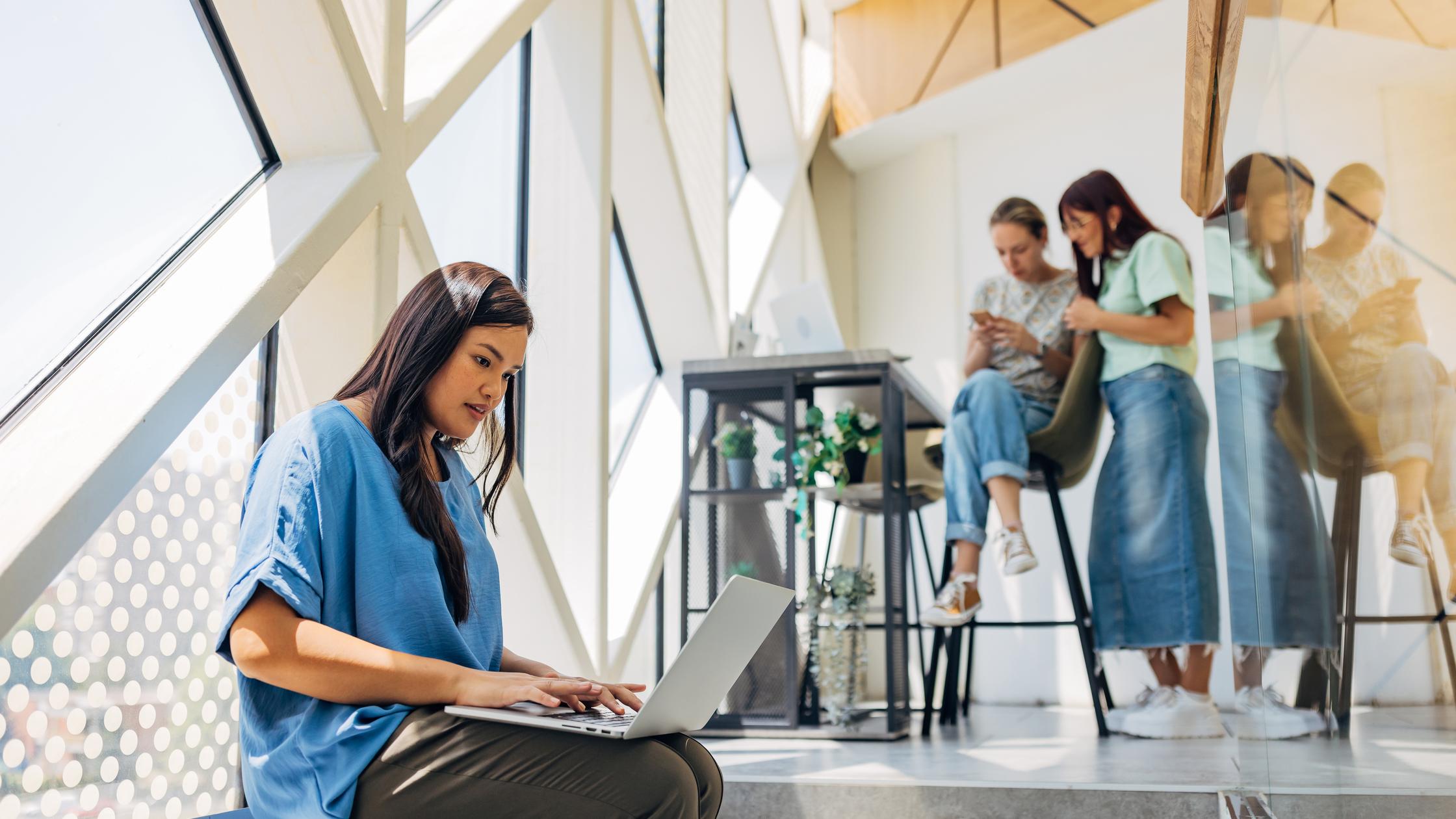 Business woman sits not far from her colleagues in a hybrid business environment and uses a laptop