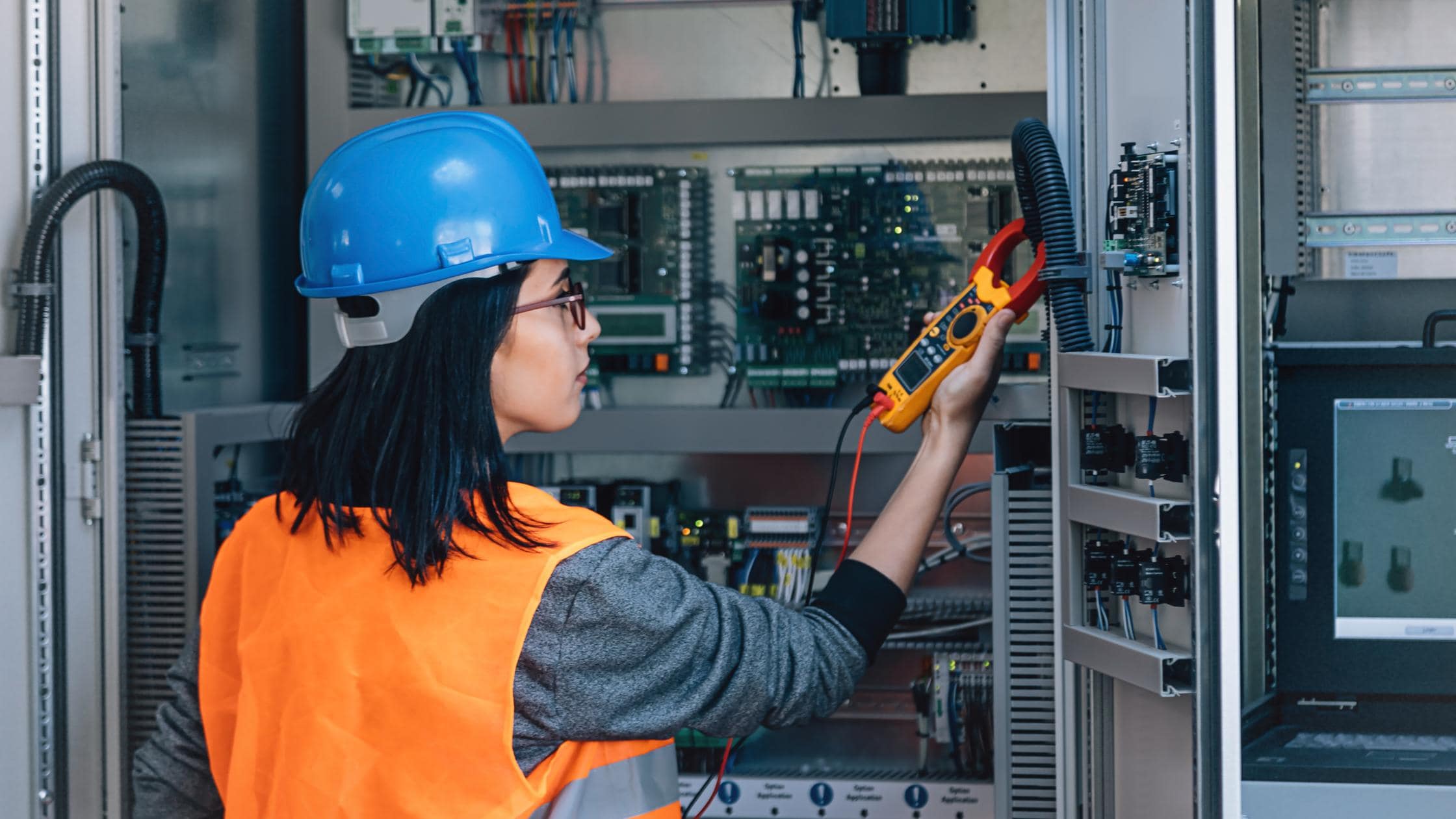 Young woman industrial service electrician engineer wearing protective vest and blue technician helmet testing and checking fridge electric voltage.